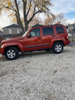 2009 Jeep Liberty in burnt orange, excellent condition, parked on gravel with trees in background.