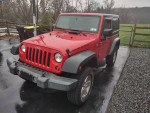 2009 Jeep Wrangler in red, featuring a soft top and minor dings, parked on a wet driveway.