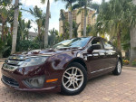 2012 Ford Fusion in raspberry red parked among palm trees, featuring polished aluminum wheels and clean interior.