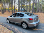 2012 Mazda 3 i Grand Touring sedan parked on a forest road, showcasing its sleek design and shiny exterior.