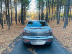 2012 Mazda 3 i Grand Touring parked on a road surrounded by pine trees, showcasing its smooth lines and design.