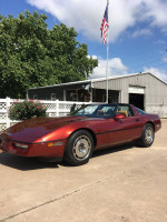 1987 Chevrolet Corvette in red color parked outdoors with a flag and green trees.