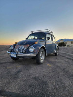 1970 VW Beetle with patina, new seats from a 2015 Kia Soul, parked on asphalt at sunset.